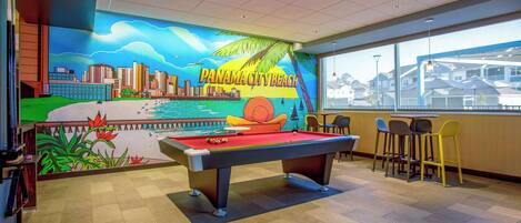A pool table with a mural of Panama City Beach, a bar area with chairs, and a view of houses through large windows.