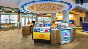 A modern hotel lobby with a central counter displaying beverages and snacks, large windows offering views of the outdoors, and a seating area with wooden tables and chairs. - Tru By Hilton Panama City Beach (Panama City Beach)