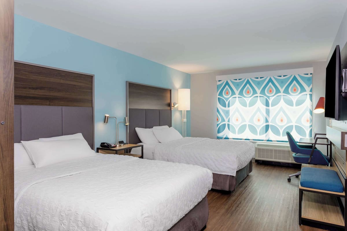 A hotel room with two beds, a desk, a chair, and a TV.
