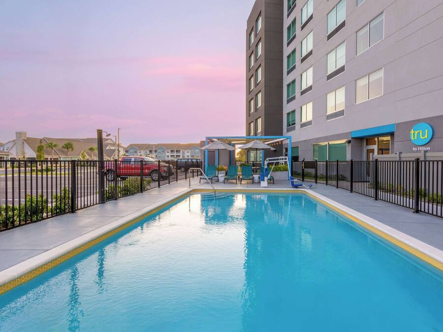 A swimming pool area with lounge chairs and umbrellas, surrounded by a fence, and a hotel building in the background.