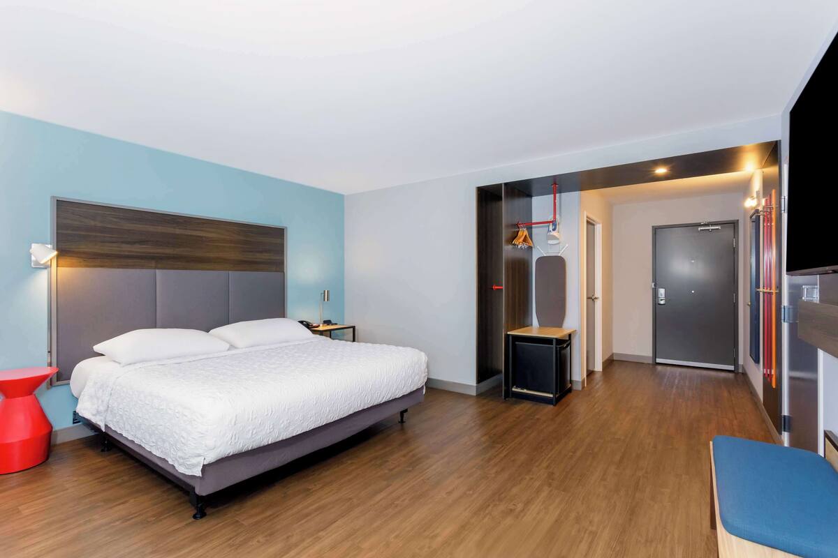 A modern hotel room with a large bed, a red side table, a small desk, and a wardrobe.