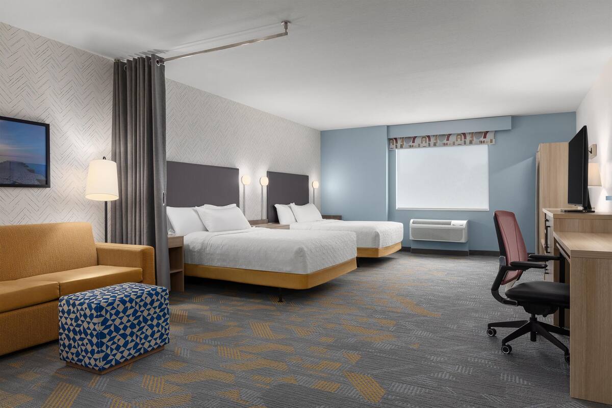 Une chambre d’hôtel avec deux lits, un canapé, un bureau et une chaise. Il y a une fenêtre avec des rideaux, un téléviseur fixé au mur et une lampe.