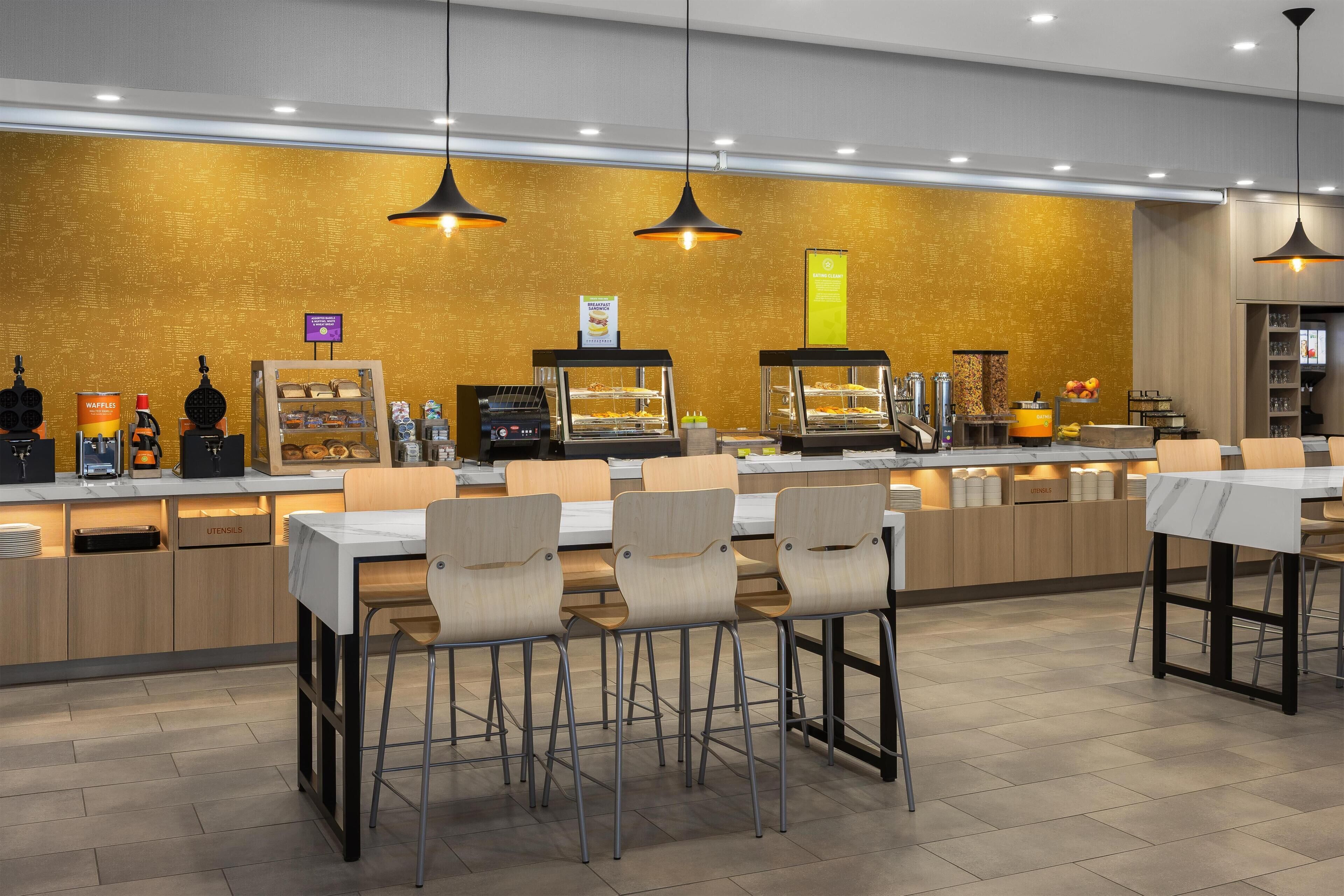 Un café moderno con mostrador exhibiendo diferentes productos alimenticios, zona de estar con mesas y sillas, e iluminación colgante.