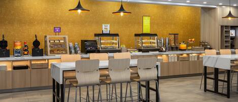 Un café moderno con mostrador exhibiendo diferentes productos alimenticios, zona de estar con mesas y sillas, e iluminación colgante.