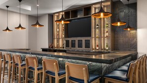 Zona de bar con sillas de madera, una barra de mármol, iluminación colgante y una pantalla de televisión grande.