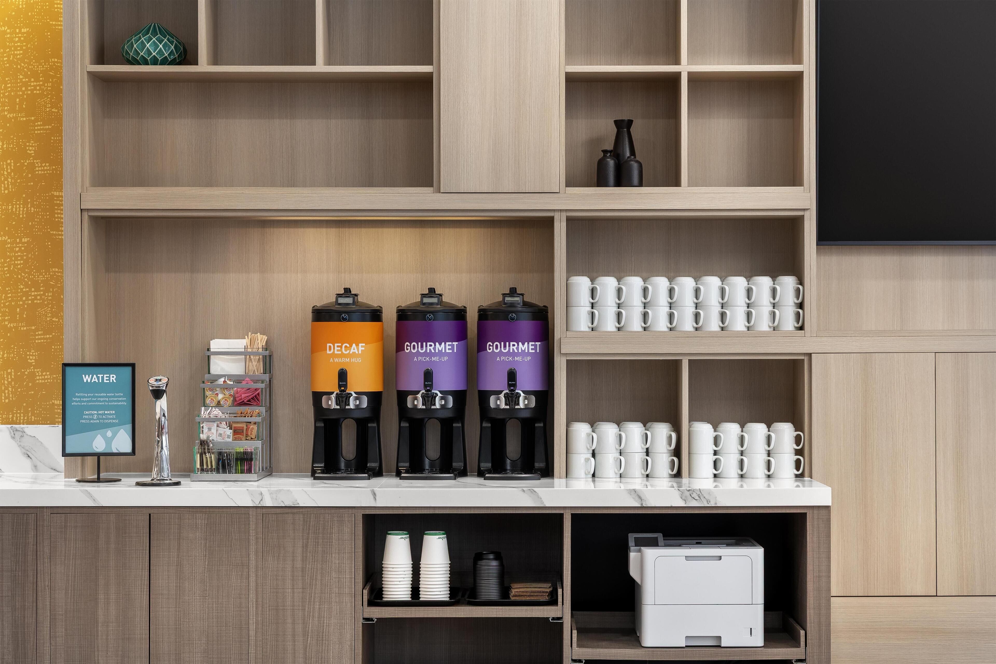 A coffee station with three coffee makers labeled 'Decaf', 'Gourmet', and 'Gourmet'. There are stacks of white cups and a water dispenser.