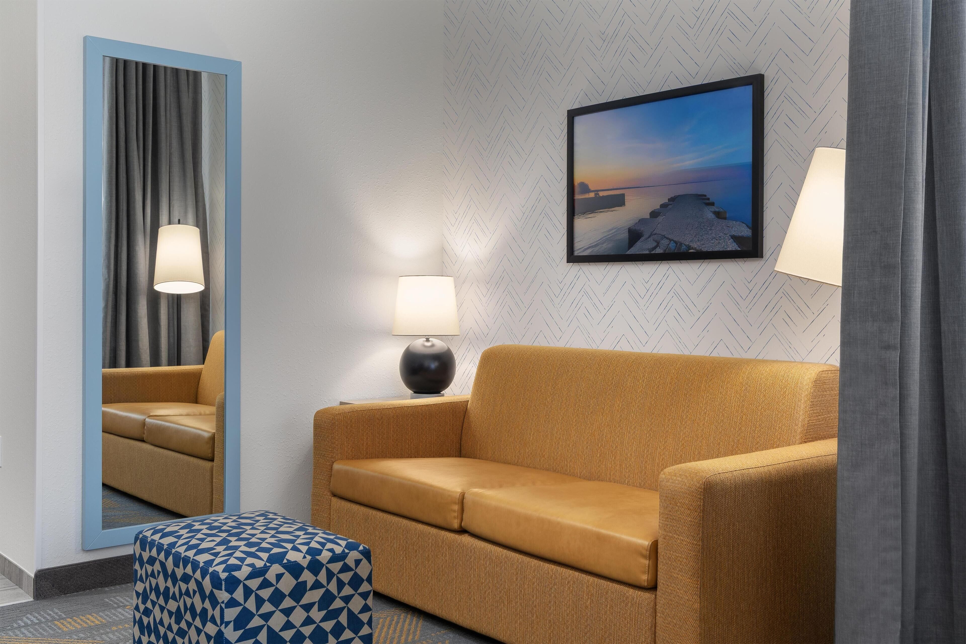 A hotel room with a sofa, a patterned ottoman, a mirror, a lamp, and a framed picture on the wall.