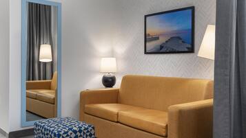 A hotel room with a sofa, a patterned ottoman, a mirror, a lamp, and a framed picture on the wall.