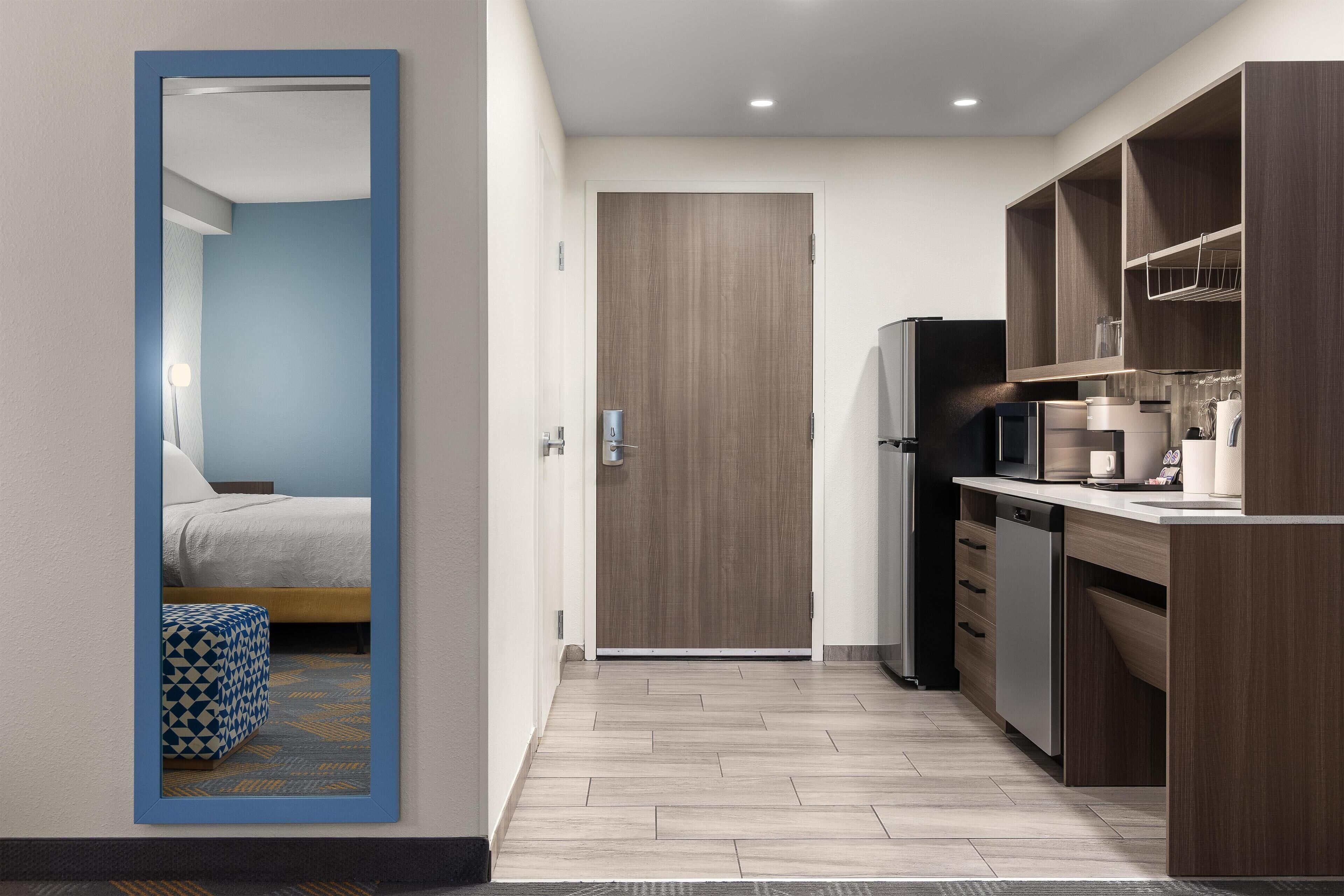 Een moderne keuken met een magnetron, koelkast en houten kastjes. Een deur leidt naar een slaapkamer met een bed en een gestreept poef.
