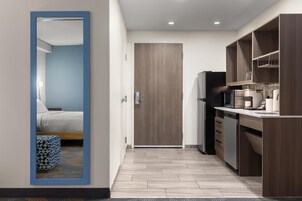 Cozinha moderna com micro-ondas, frigorífico e armários de madeira. Uma porta dá acesso a um quarto com cama e um banquinho estampado.