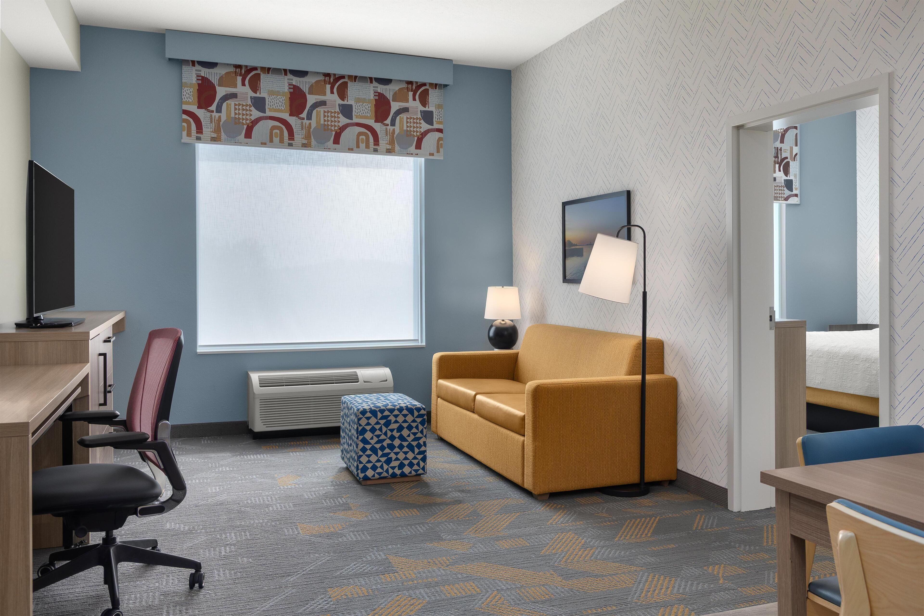 A hotel room with a desk, office chair, sofa, ottoman, lamp, and a bed with a headboard.