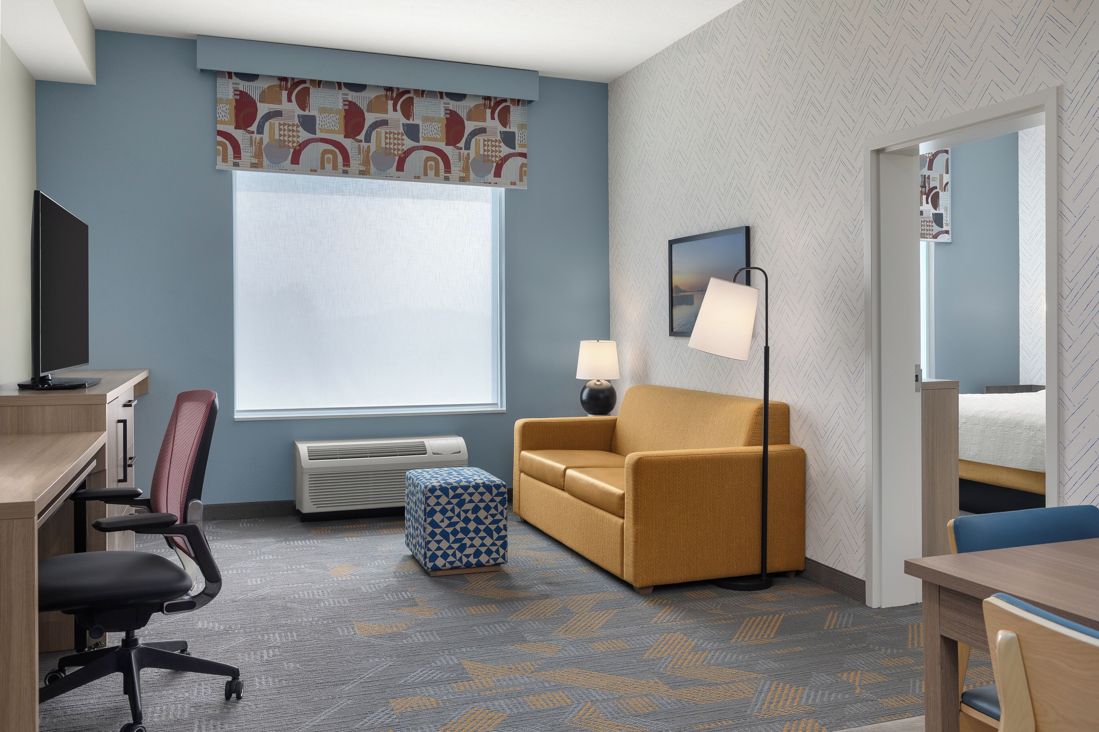Una habitación de hotel con escritorio, silla de oficina, sofá, otoman y cama con cabecera.