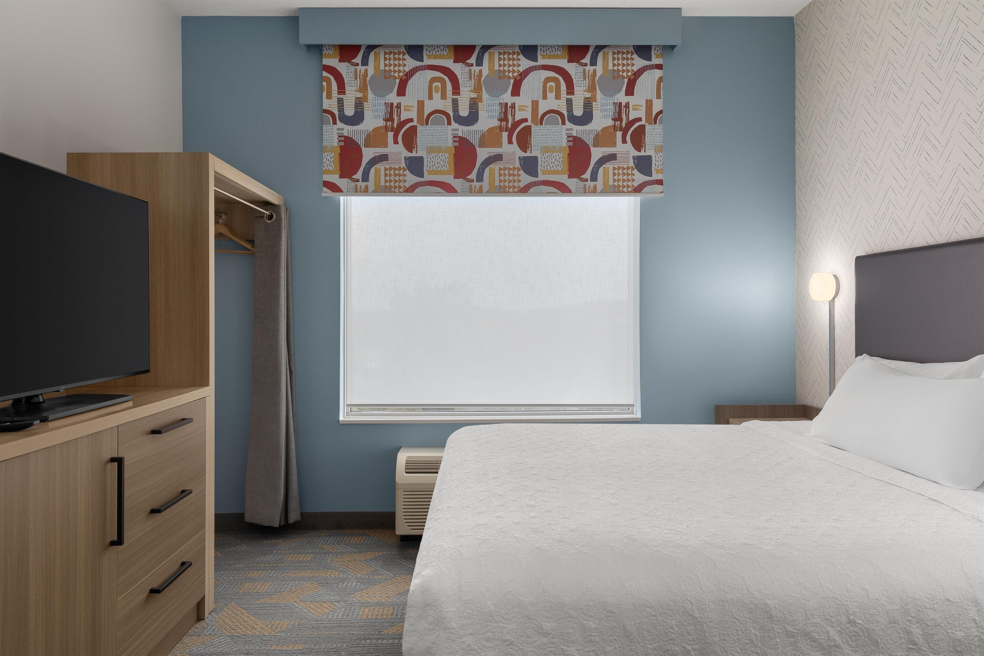 A hotel room with a bed, a TV on a wooden dresser, a window with patterned curtains, and a nightstand with a lamp.