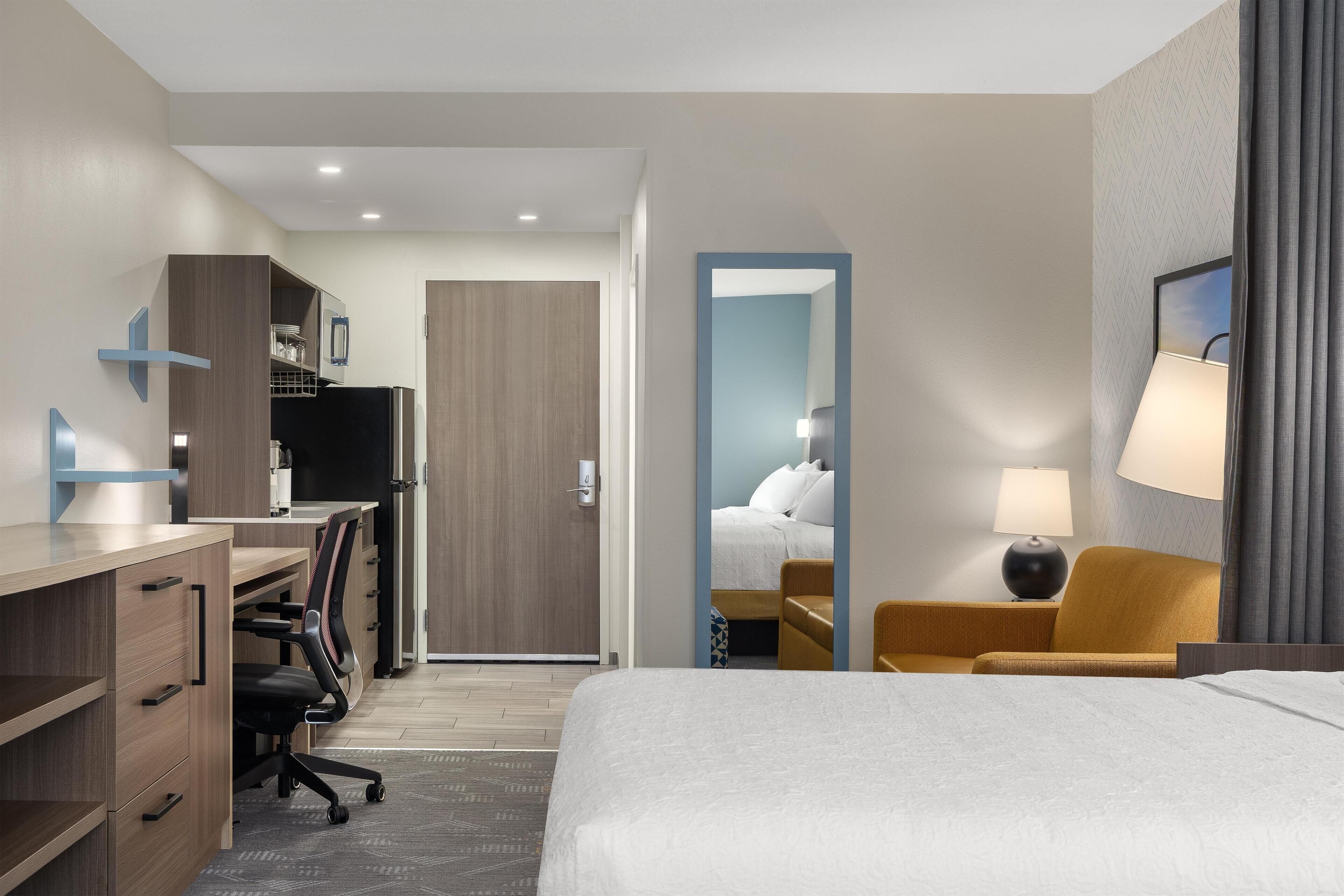 A hotel room with a bed, desk, chair, and a view into another room.