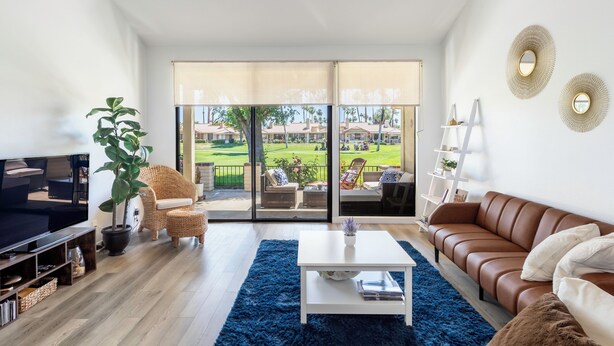 Una sala de estar moderna con un televisor grande, asientos cómodos, una mesa de centro y vistas a un área exterior verde a través de puertas de vidrio.