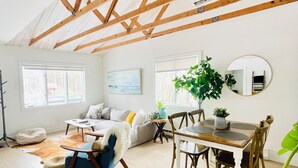 A spacious living room with a high ceiling, wooden beams, a dining area with a table and chairs, a sofa, and a large window.