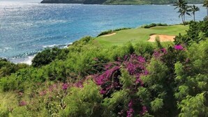 A coastal landscape with a golf course, lush greenery, and a clear view of the ocean and distant mountains.