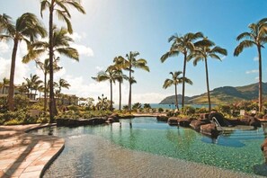 Une piscine d’un complexe hôtelier, bordée de palmiers, avec un ciel dégagé et une chaîne de montagnes en arrière-plan.