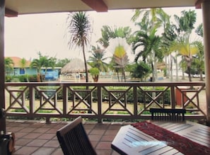 Un espacio al aire libre cubierto con barandilla de madera, un banco y una mesa con mantel rojo. Palmeras y una estructura con techo de paja se observan al fondo.