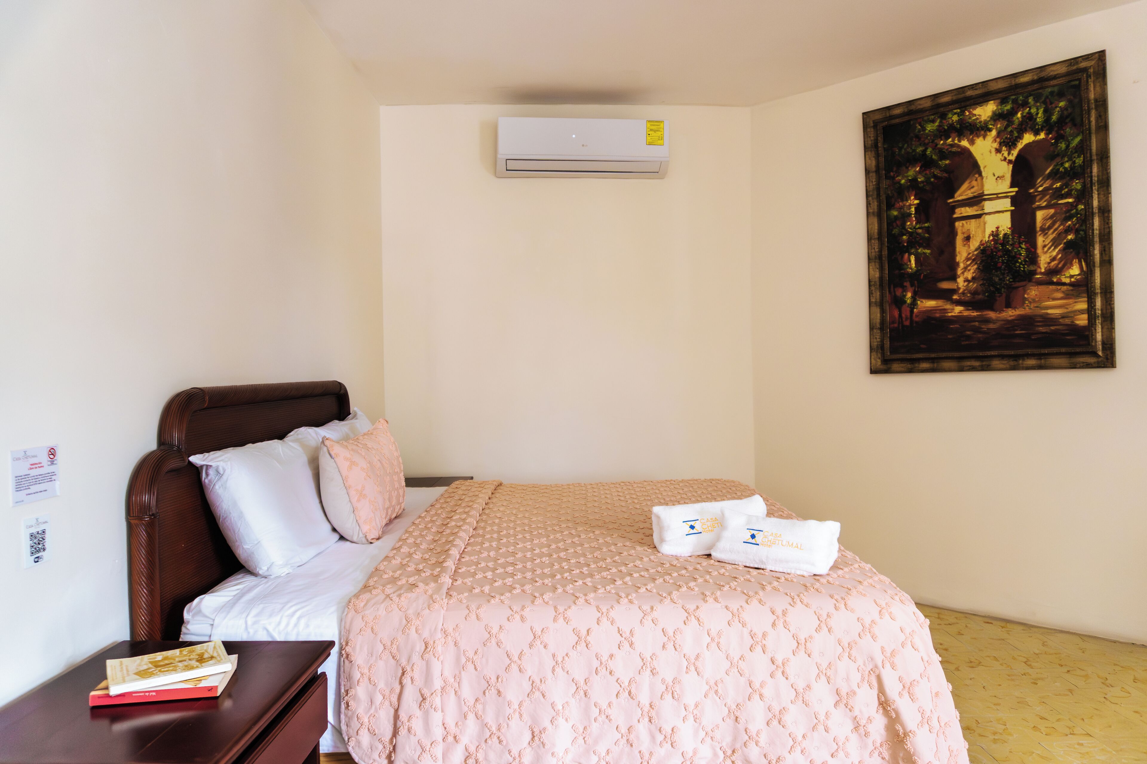 A neatly made bed with a pink patterned bedspread, a wooden headboard, and two towels on the bed. There is a small table with a book and a framed picture on the wall.
