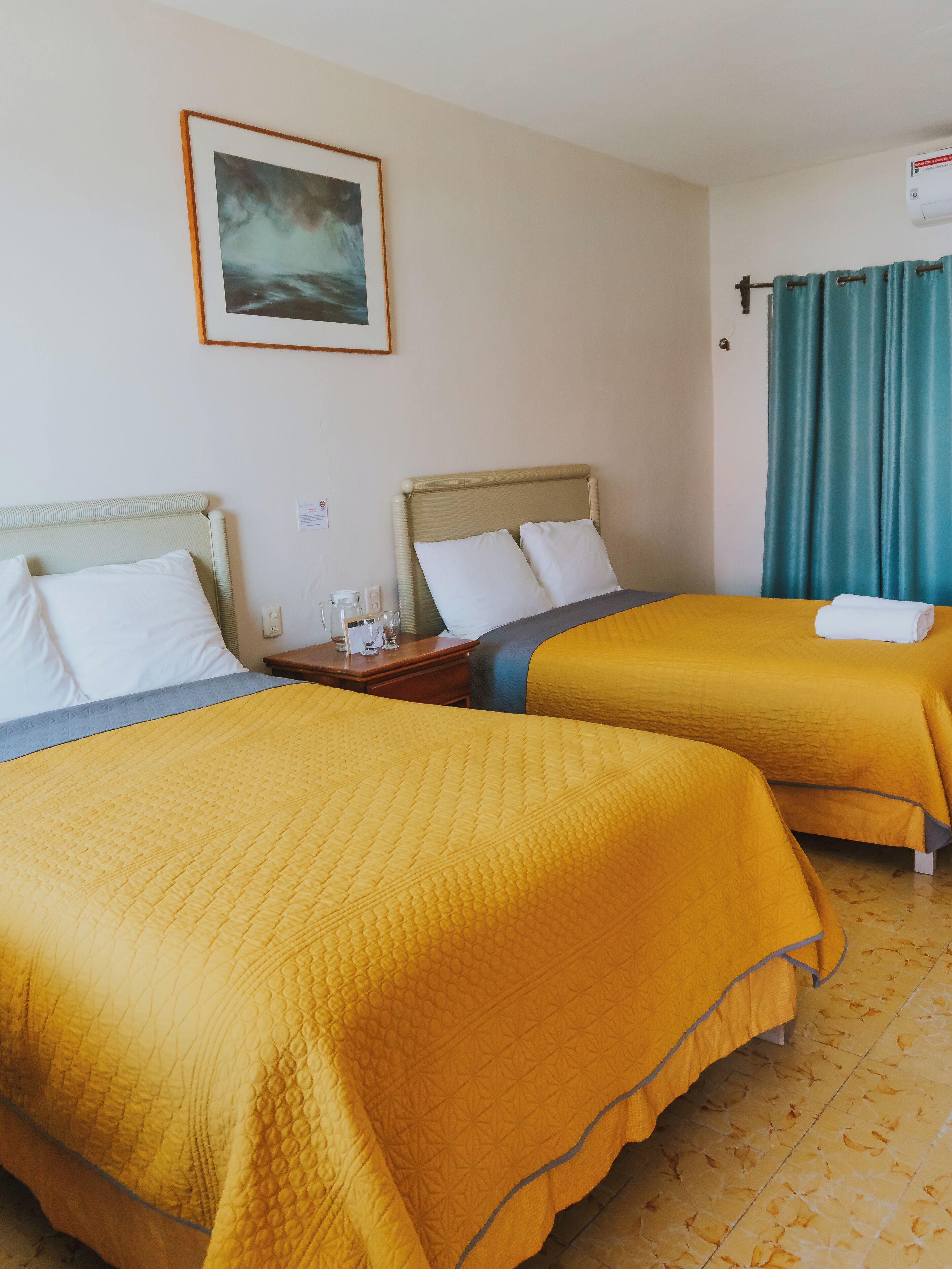 Habitación de hotel con dos camas, cada una con cubrecama amarillo, un cuadro en la pared y una ventana con cortinas color turquesa.