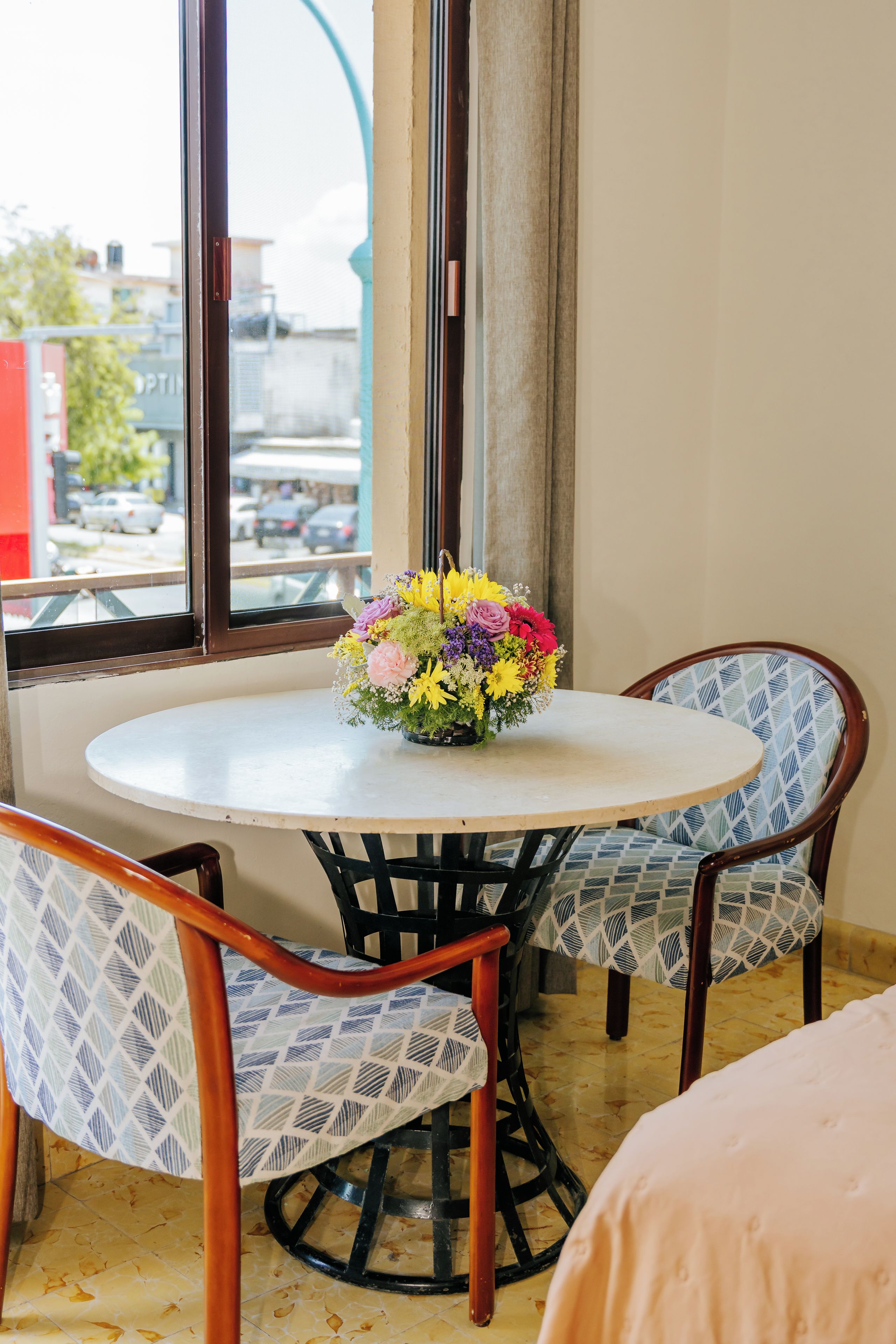 Una mesa redonda con un centro de mesa floral, dos sillas estampadas y vista a la calle a través de una ventana.