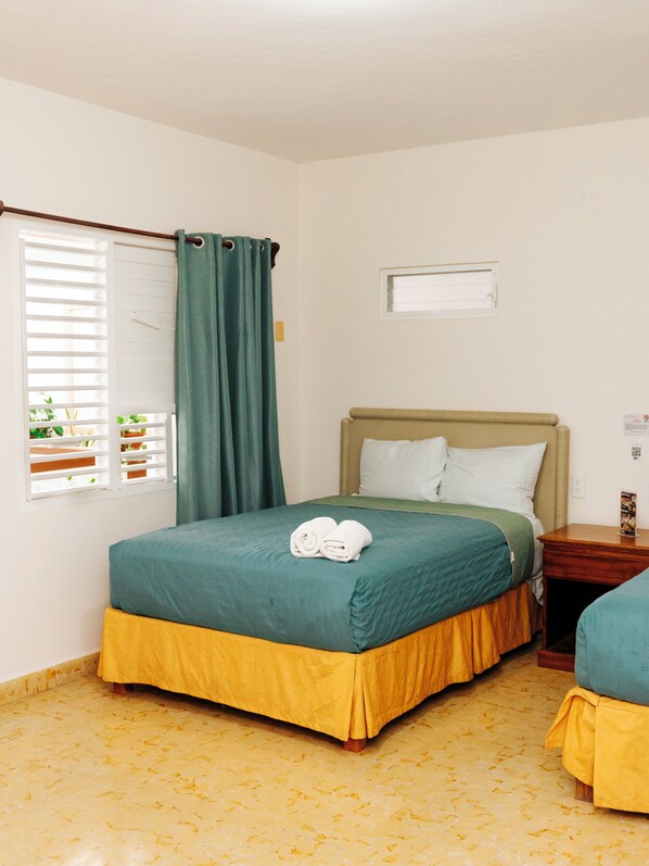 A single bed with a green and yellow bedspread, a wooden nightstand with a lamp, and a window with blinds and a curtain.