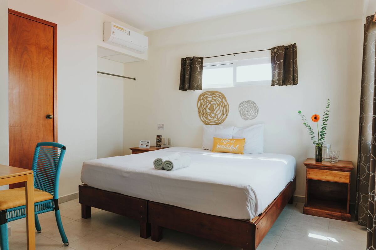 A hotel room with a wooden bed, a wooden nightstand, a blue chair, a small table, and a window with curtains.