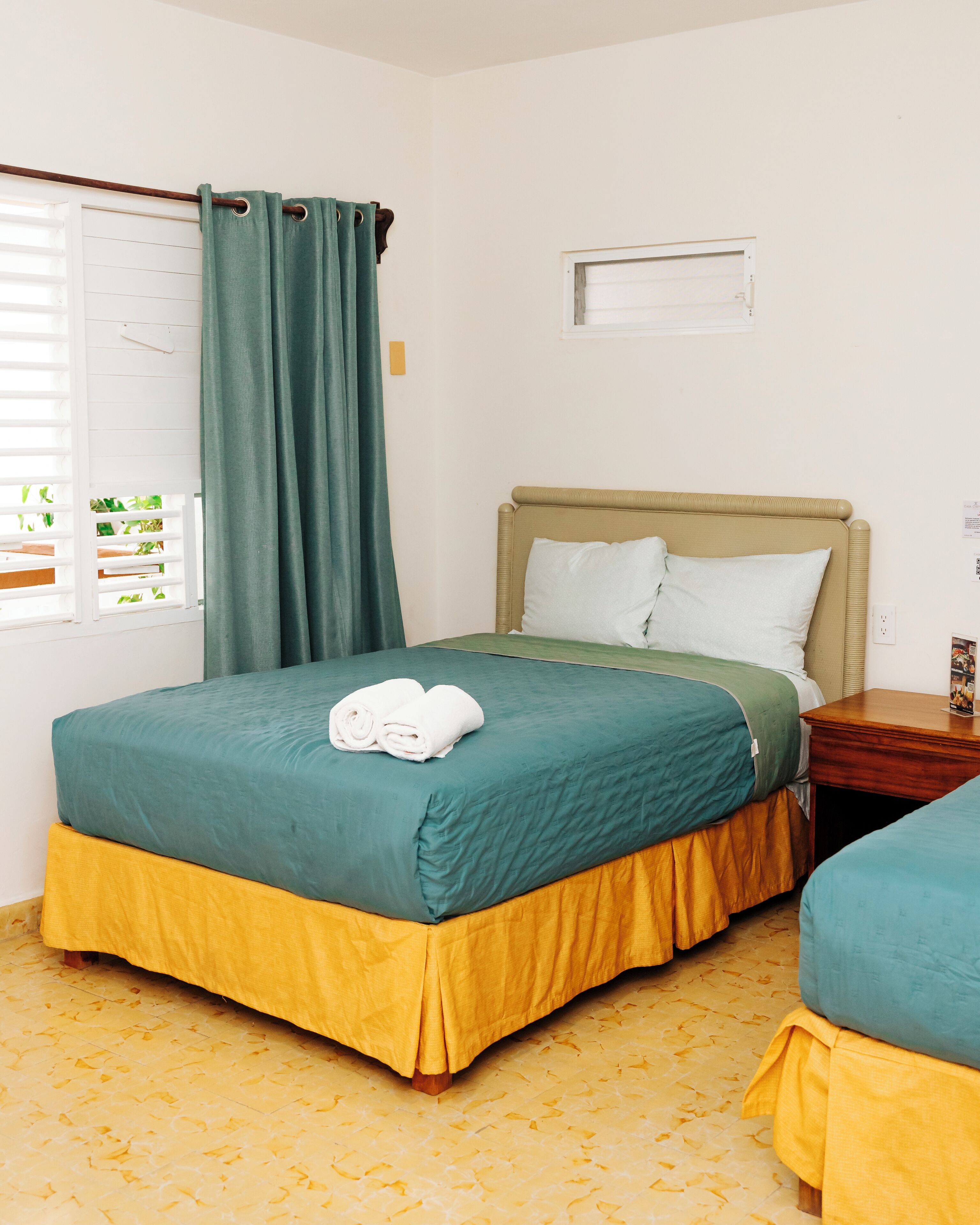 Una cama individual con una colcha verde y amarilla, una mesita de noche de madera, una ventana con persianas blancas y una cortina.