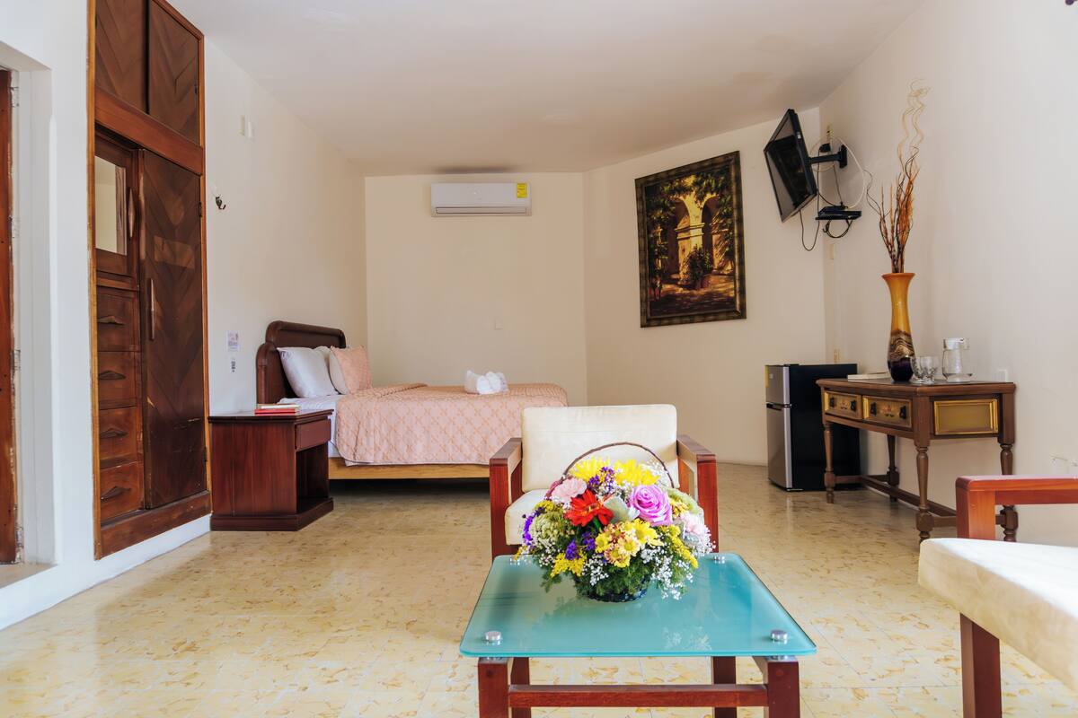 A bedroom with a bed, a wooden wardrobe, a small table with a vase of flowers, a chair, a desk, and a television mounted on the wall.