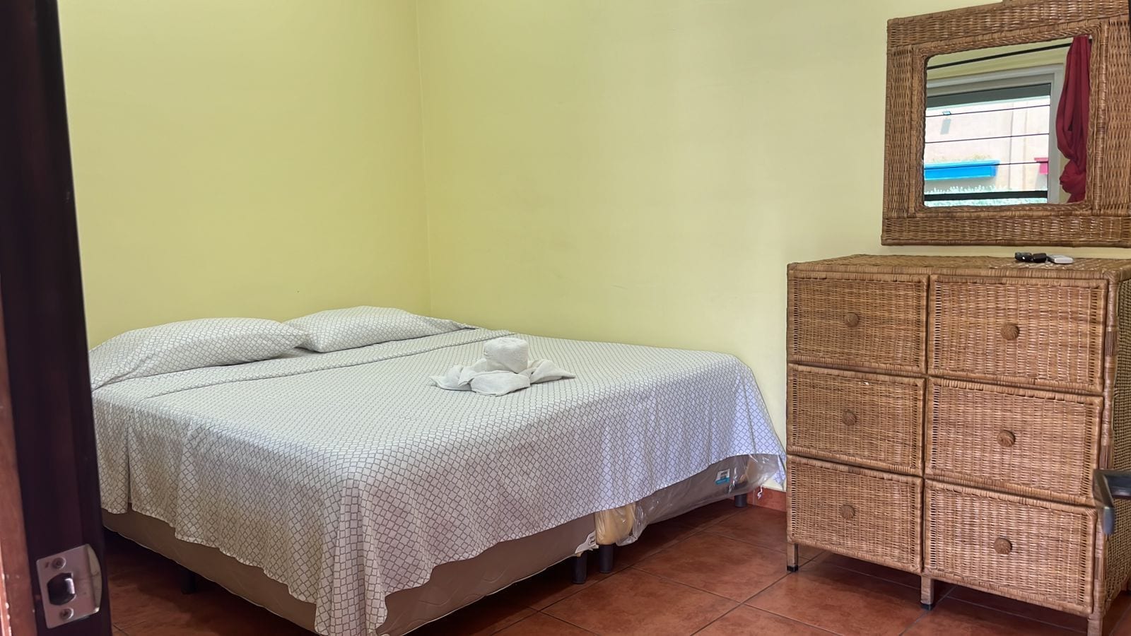 Un llit individual amb un estovat i coixins estampats, un moble d'emmagatzematge de vimetla i un mirall amb vista a l'exterior.