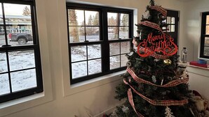 Ein Zimmer mit einem Weihnachtsbaum, geschmückt mit karierten Bändern und einem Schild mit der Aufschrift 'Merry Christmas'. Drei Fenster bieten Blick auf eine verschneite Landschaft.