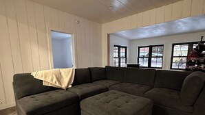 Ein Wohnzimmer mit einer Eckcouch, einem Hocker, Holzwandverkleidung und großen Fenstern.