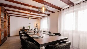 A dining area with a long wooden table set for six, black leather chairs, wooden beams on the ceiling, and a hanging pendant light. - The Cottage (Guémar)