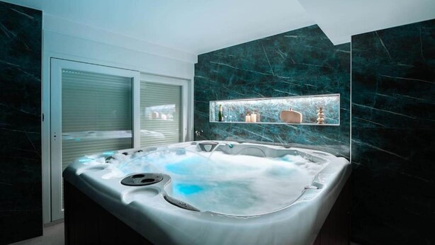 A modern bathroom with a spa tub, marble walls, and a window with blinds. - The Cottage (Guémar)