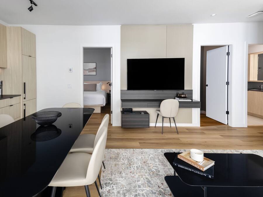 Ett modernt kök med en svart bänkskiva, en matplats med ett svart bord och stolar, en platt-TV som är monterad ovanför ett skrivbord och ett litet pentry.