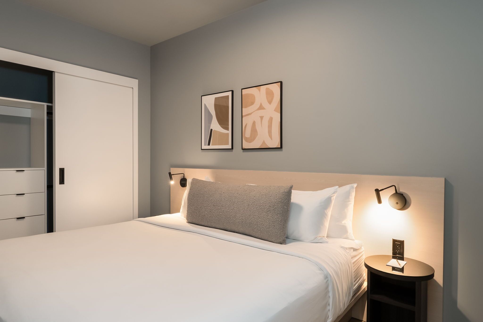 Ein ordentlich bezogenes Bett mit Kopfteil, ein Nachttisch mit Lampe und zwei gerahmte Kunstwerke an der Wand.