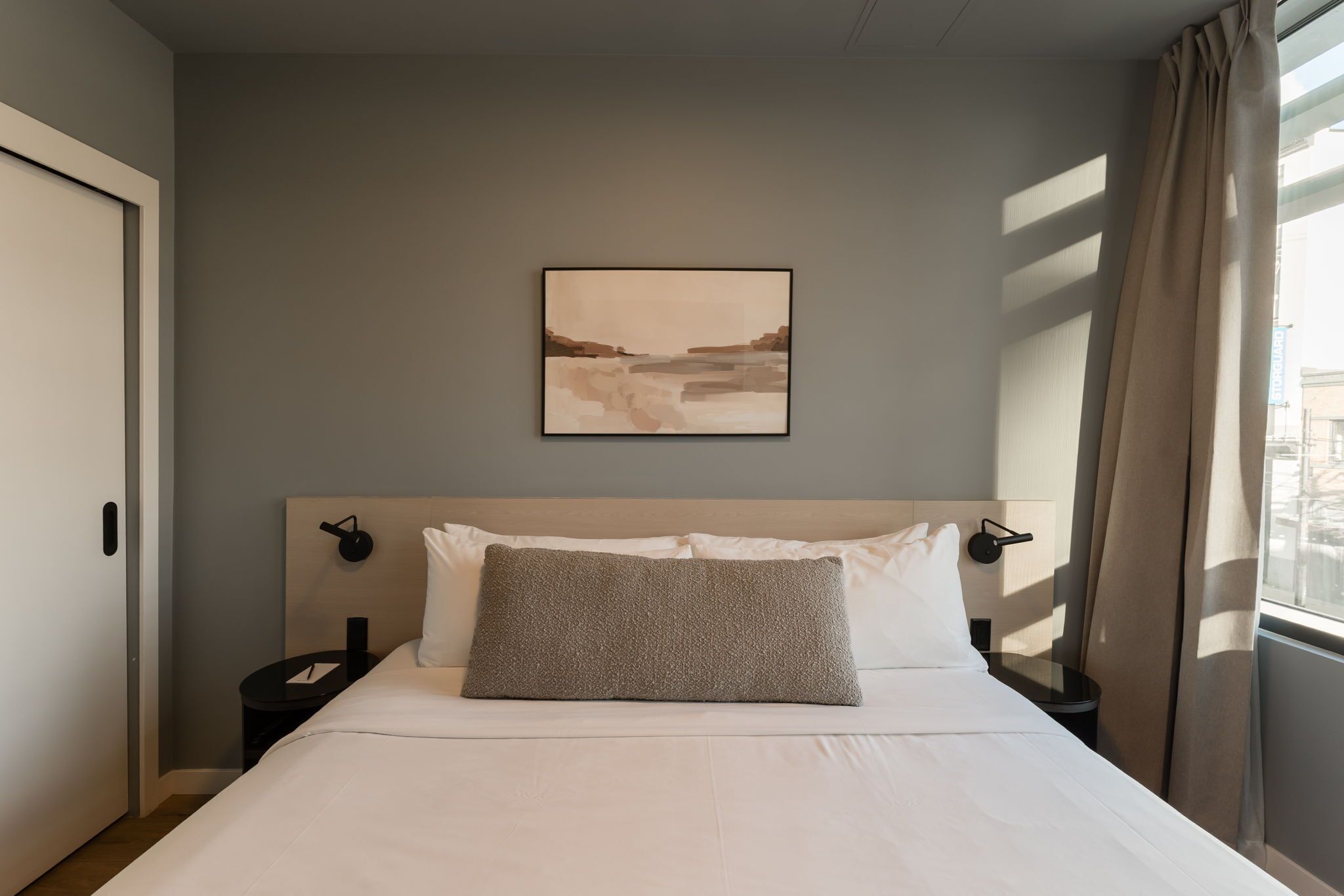 A hotel room with a bed, a nightstand, a wall-mounted lamp, and a window with curtains.