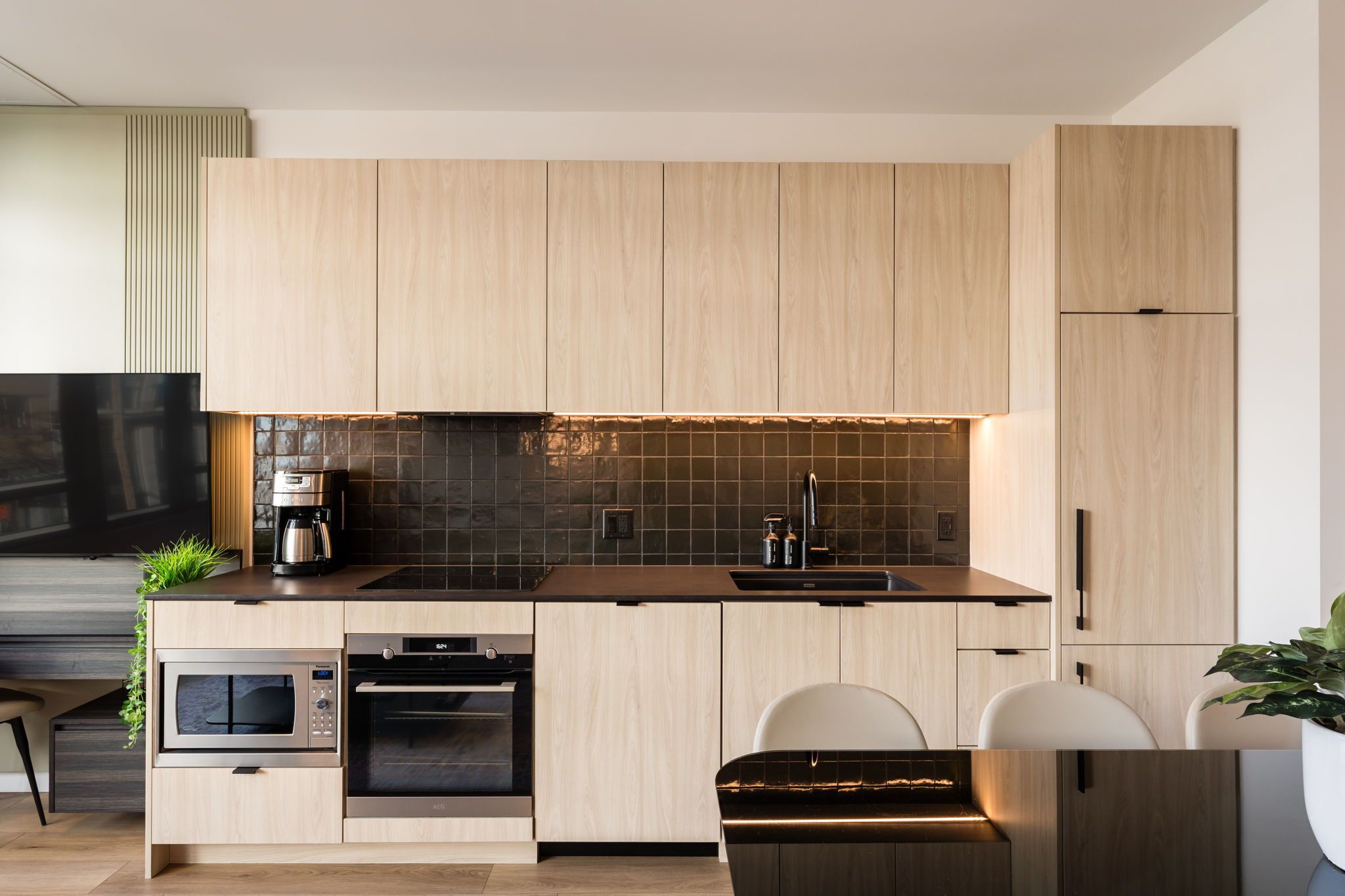 Une cuisine moderne avec des meubles en bois clair, un dosseret en carreaux noirs, des appareils en acier inoxydable et un four encastré.