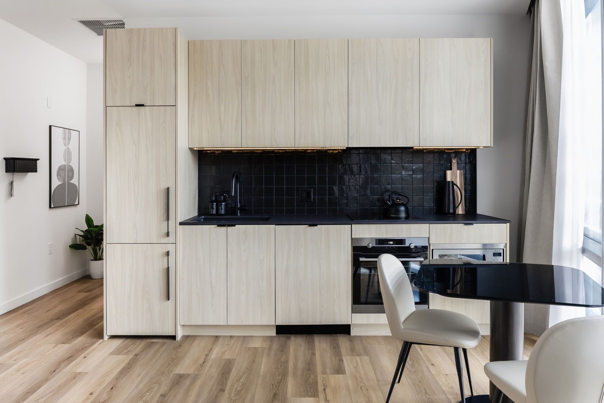 A modern kitchen with light wood cabinets, a black countertop, and a dining area with a glass table and white chairs.