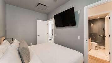 A modern bedroom with a bed, a flat-screen TV mounted on the wall, and a visible bathroom with a glass shower enclosure.