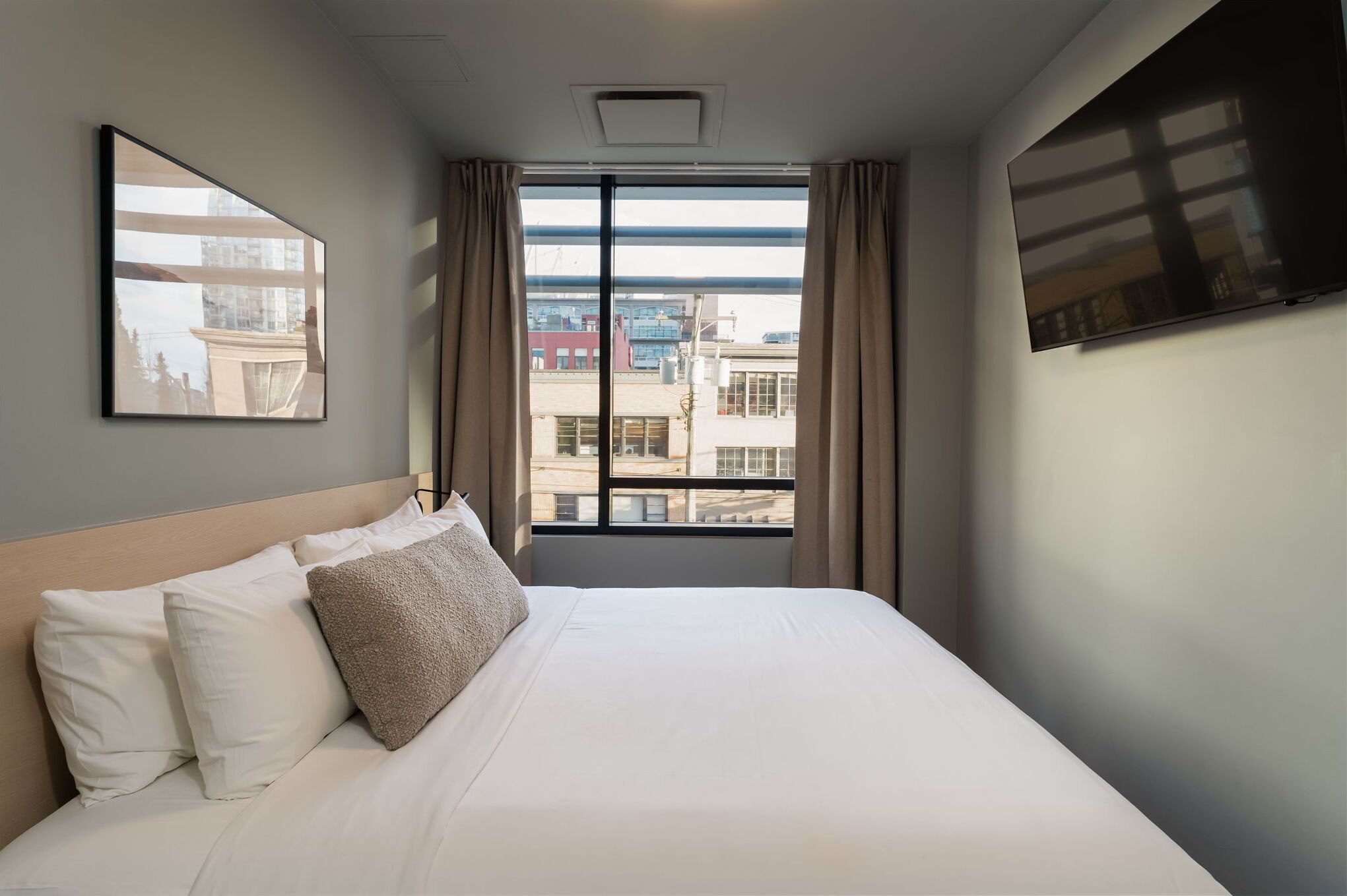 A hotel room with a large bed, a view of buildings through the window, and a mounted TV on the wall.