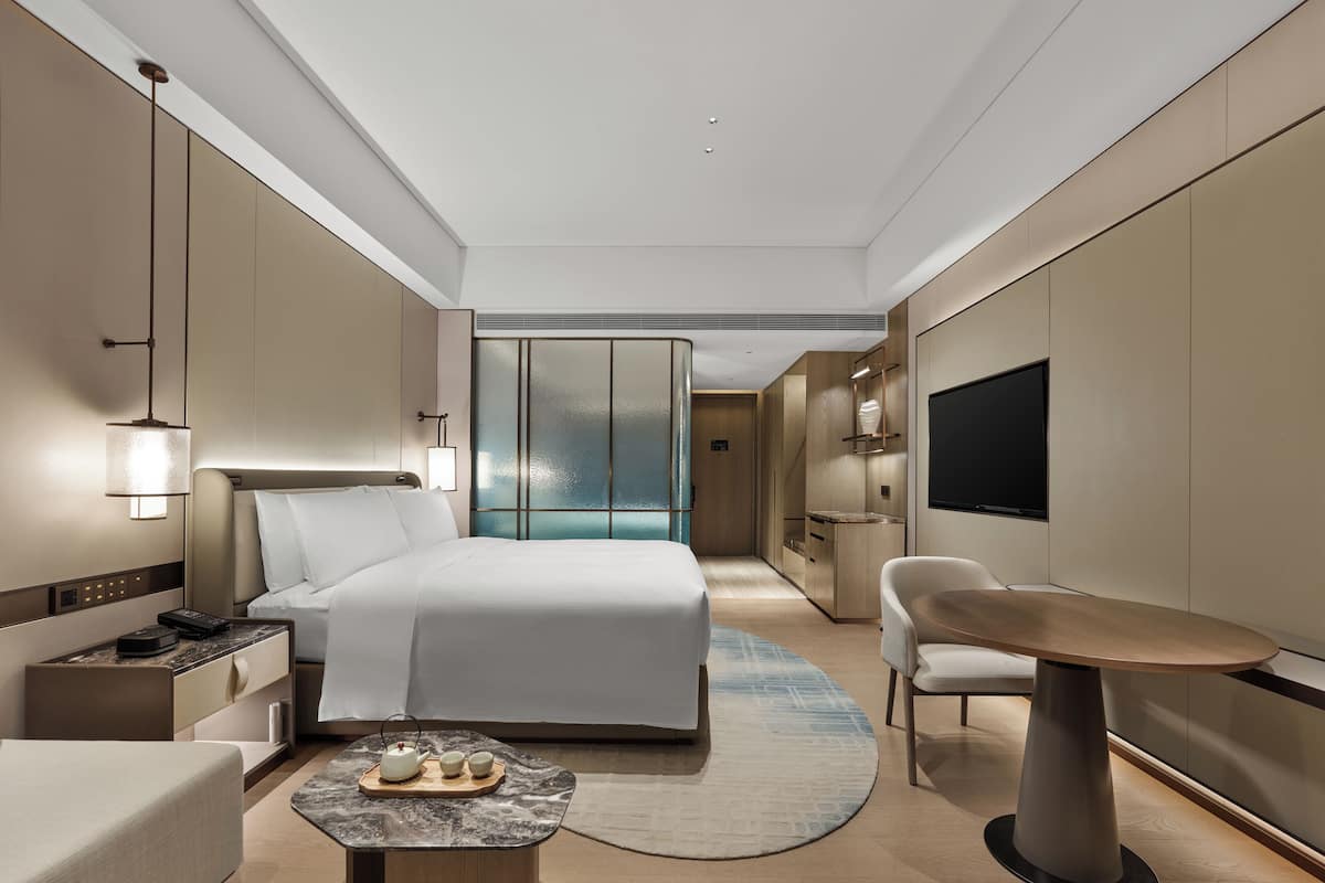 Quarto de hotel moderno com cama, mesa de cabeceira, televisão e um espaço de estar com mesa e cadeiras.