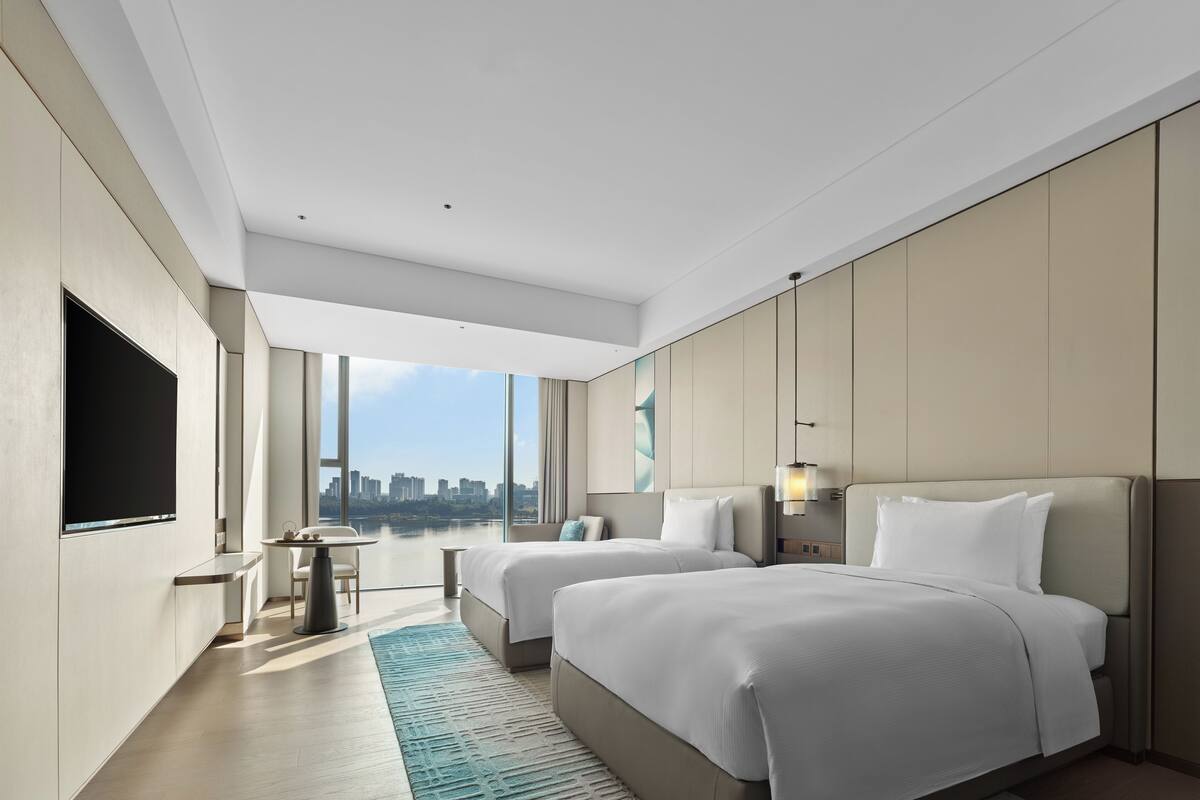 Quarto de hotel moderno com duas camas, televisão, vista para a cidade e uma pequena mesa.