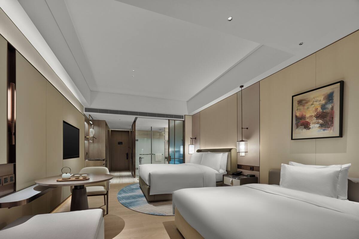 Quarto de hotel com cama, escrivaninha, cadeira e banheiro visível através de uma divisória de vidro.