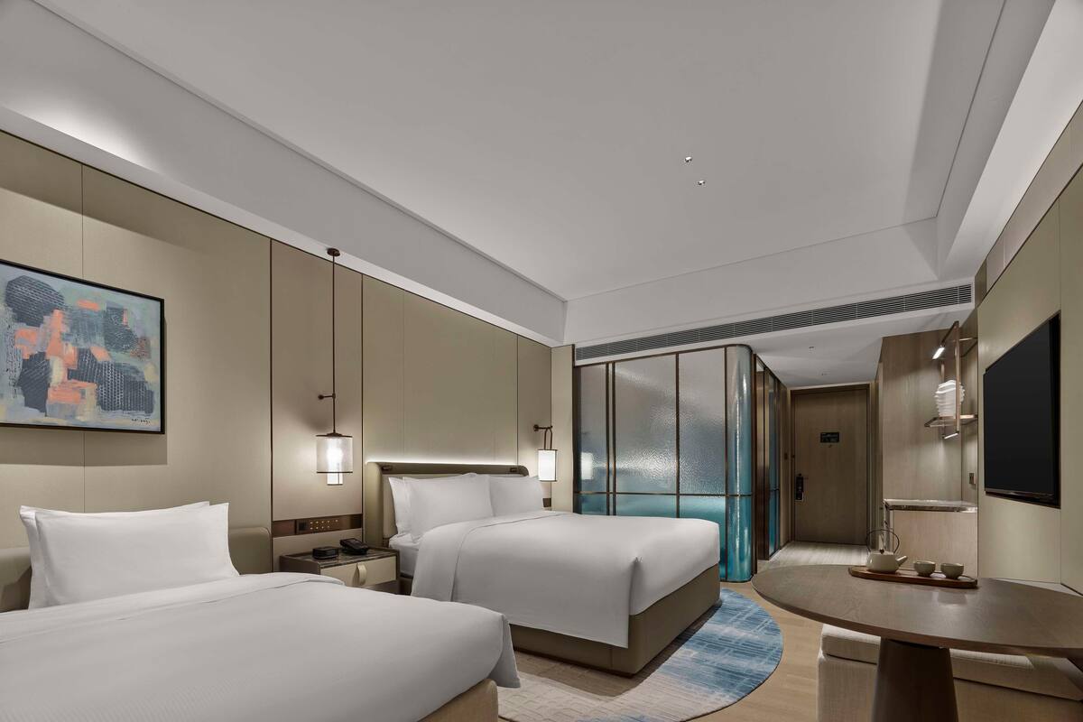 Quarto de hotel com duas camas, uma escrivaninha, televisão e um quadro na parede.
