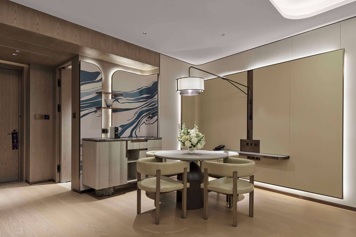Quarto de hotel moderno com área de refeições, apresentando uma mesa redonda e quatro cadeiras, um grande quadro abstrato na parede e um design minimalista em tons de madeira e cores neutras.