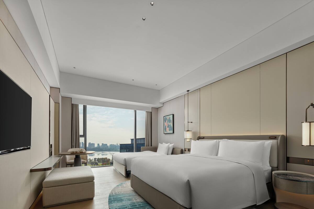Quarto de hotel moderno com uma cama grande, área de estar com mesa, TV e vista para a cidade.