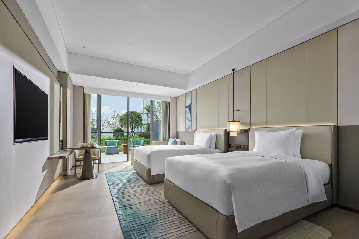 Quarto de hotel moderno com duas camas, televisão de tela plana, uma mesinha e um espaço de estar com cadeiras.