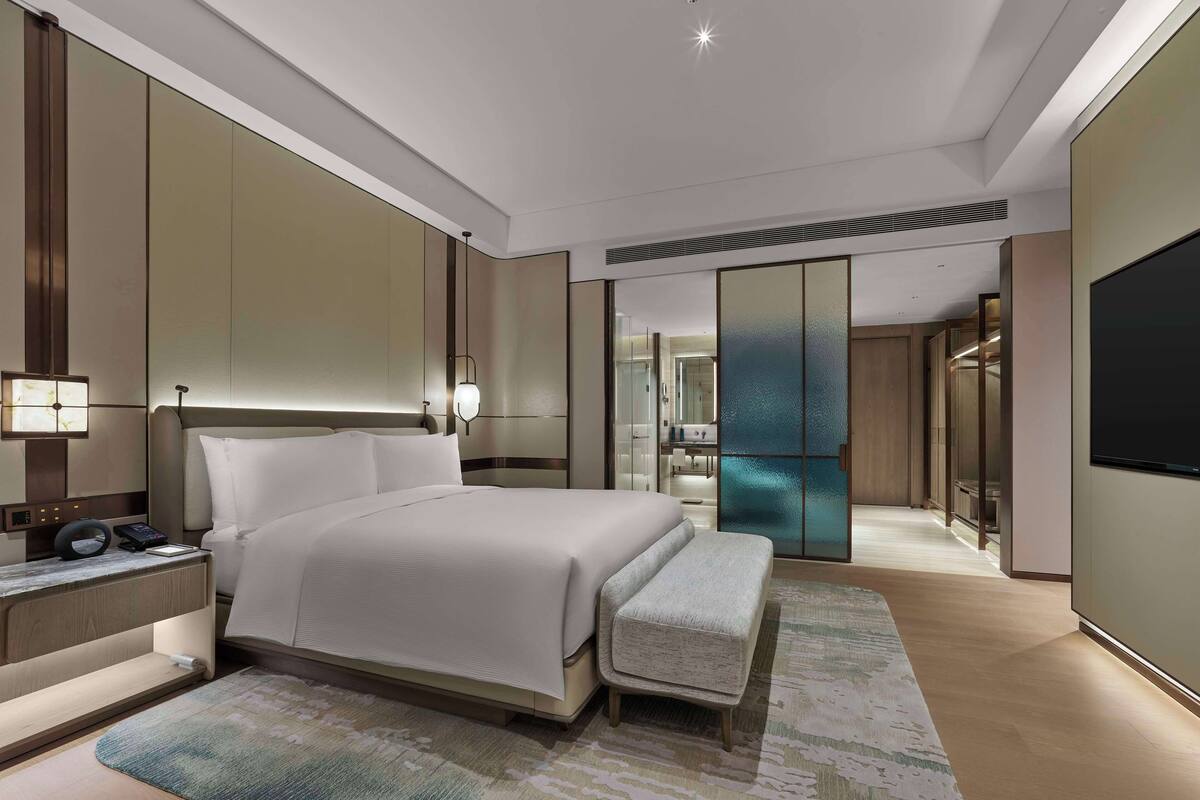 Quarto de hotel moderno com uma cama grande, mesa de cabeceira, luminária e um banco.